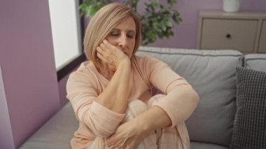 Olgun, sarışın, beyaz, kadın oturma odasında yalnız oturuyor, düşünceli ve düşünceli görünüyor, kapalı bir evde, rahat bir kanepede, arka planda bir bitki ve dolap ile..