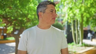 Şehir parkında arka planda yeşillik olan genç bir adam dalgın dalgın bakıyor ve beyaz bir gömlek giyiyor.