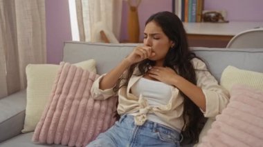 Kanepede pembe yastıklarla rahat bir oturma odasında öksüren, sıkıntılı bir yüz ifadesi ve rahat bir kıyafet sergileyen, ev ortamında kendini kötü hisseden bir kadın.