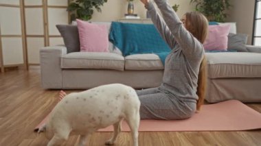 Genç bir kadın evindeki evcil köpeğiyle yoga minderinde egzersiz yapıyor. Etrafı modern mobilyalar ve rahat dekorlarla çevrili..