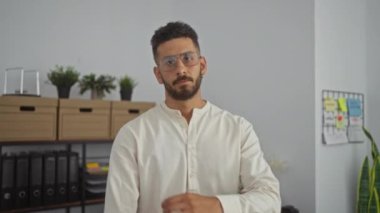 Yakışıklı, gözlüklü ve beyaz gömlekli bir adam. Kapalı bir ofis ortamında kolları çapraz duruyor. Arka planda saksı bitkileri ve ofis malzemeleri var..