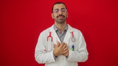 Göğsünde elleri olan, doktor üniformalı gülümseyen İspanyol genç adam, izole edilmiş kırmızı arka planda sağlığa karşı minnet dolu bir jest gösteriyor.