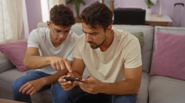 Baba ve oğul renkli bir salonda birlikte oturuyorlar, evde kaynaşırken akıllı bir telefona odaklanıyorlar.