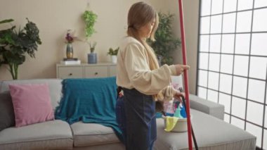 Genç bir kadın, evcil köpeğinin yanında paspasla oturma odasını temizliyor. Evinin içi bitki ve modern dekorlarla dolu..