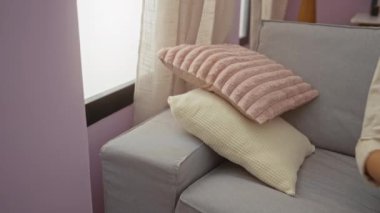 Gri bir kanepede, bej perdeleri ve pastel duvarları olan rahat bir oturma odasında, yumuşak pembe bir yastık tutan bir kadın.