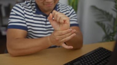 Genç Çinli bir adam bir ofiste oturuyor, hafif bir gülümsemeyle bileğine masaj yapıyor, masaüstü bilgisayarı kullanırken çizgili bir gömlek giyiyor..