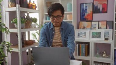 Arka planda bitkiler ve sanat eserleri olan bir ev dekorasyonu mağazasında dizüstü bilgisayarda çalışan genç bir adam, çevrimiçi etkileşim halindeyken çeşitli duyguları gösteriyor