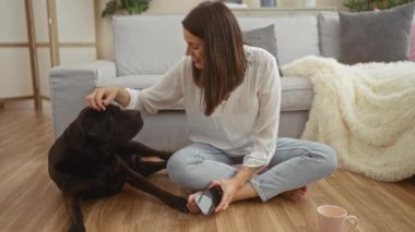 Bir kadın, elinde akıllı bir telefon tutarken labrador köpeğini okşayarak oturma odasında yerde oturuyor..