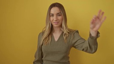 Sarışın kadın, el pençe divan duruyor. Parmaklarının arasından bakıp gülümsüyor. Sarı arka planda soyutlanmış.