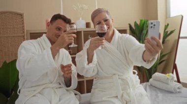 Bornozlu kadın ve erkek şarap içip spa odasında selfie çekiyorlar. Rahatlatıcı bir ev ortamı sergiliyorlar.