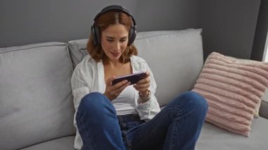 Oturma odasında kulaklıkla oynayan genç bir kadın, koltukta pembe yastıkla rahatlatıcı bir anın tadını çıkarıyor.