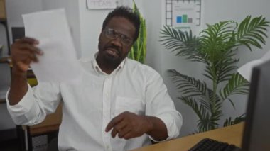 Yakışıklı, genç, Afro-Amerikalı bir adam kapalı ofis ortamında belgelerle serinliyor. Etrafı fabrikalar ve ofis mobilyalarıyla çevrili..
