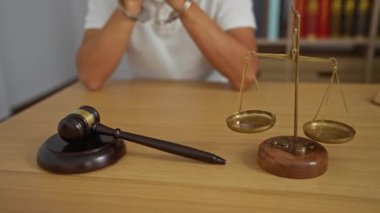 Elleri kelepçeli genç bir adam, bir hukuk bürosunun ahşap masasında oturuyor. Arka planda hukuk kitapları, yargıç tokmağı ve denge terazilerinin yanında.