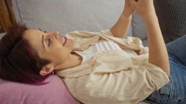 Evdeki bir kanepede dinlenen kısa saçlı genç ve güzel bir kadın. Evinin oturma odasındaki akıllı telefonu kullanıyor..