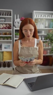 Stresli kadın iç mekan dekorasyon dükkanında para sayıyor. Yanında dizüstü bilgisayarı ve not defteri var. Mali sıkıntıları ya da iş sorunlarını gösteriyor..