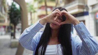Genç bir esmer kadın, şehir sokaklarında gülümserken, şehir dışında bir yerde mutluluğunu sergilerken elleriyle kalp şekli yapar..