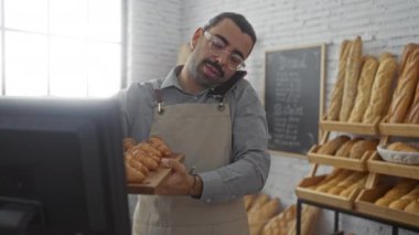 Genç adam, arka planda ekmek rafları olan bir fırında kruvasan tutarken telefonla konuşuyor.