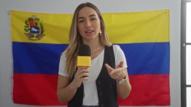 Genç, güzel, sarışın bir kadın, elinde mikrofon, Venezuela bayrağının önünde duruyor..