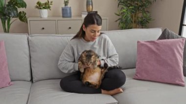 Oturma odasındaki bir kanepede rahat bir şekilde oturan genç bir kadın, elinde tekir bir kedi, saksı bitkileri ve minderlerle ev ortamına samimi bir dokunuş ekliyor..