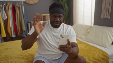 Genç bir Afrikalı Amerikalı yatak odasından canlı bir video çekiyor, bir yatağa oturuyor, akıllı telefonunu kullanıyor, neşeli bir gülümsemesi ve iç dekorasyonla çevrili sıradan bir kıyafeti var..