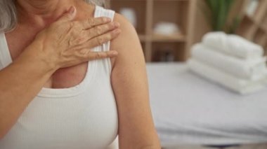 Olgun bir kadın spa odasında, eli göğsünde, beyaz bir atlet giyiyor, sağIıklı bir ortam içinde, samimi ve sakin bir kişisel bakım ve rahatlama anı yaşıyor.