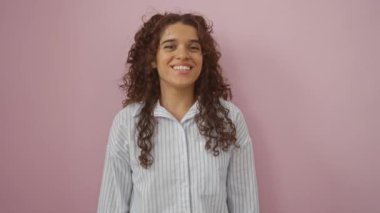 Pembe arka planda neşeli bir şekilde gülümseyen genç bir kadın kıvırcık esmer saçlarını sergiliyor ve günlük kıyafetlerini giyiyor.