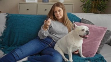 Genç bir kadın oturma odasındaki kanepede köpeğiyle birlikte rahat bir şekilde rahat bir ev elbisesi ve ev dekoru unsurları sergiliyor.. 