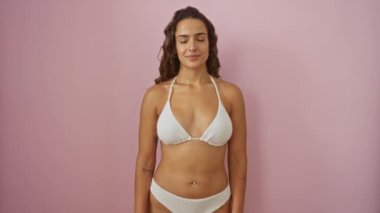 Yüzünde havalı bir gülümseme olan, pembe arka planda izole edilmiş, bikinili mutlu, genç bir İspanyol kadın.