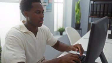 Ofisteki genç adam hayal kırıklığı içinde el ele tutuşuyor, bilgisayarla masada oturuyor, kapalı alanda bitki ve arka planda raflarla oturuyor.