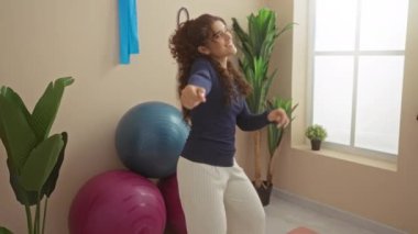 Spor salonunda dans eden bir kadın, etrafında spor topları, uzun kollu bir bluz ve pantolon giyen, bitkiler ve pencerelerle aydınlık bir ev ortamında mutluluk ve enerji ifade eden bir kadın.