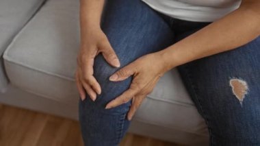 Yaşlı bir İspanyol kadın oturma odasında oturuyor ve ellerini dizlerinin üstüne nazikçe koyuyor..