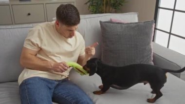 Oturma odasındaki genç İspanyol bir adam kanepede otururken daksund köpeğini uyarıyor..