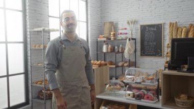 Pastanede kollarını kavuşturan yakışıklı bir adam hamur işleri ve ekmek dükkanının yanında duruyor ve karatahta menüsü olan fırınlanmış ürünleri sergiliyor..