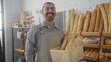 Fırında gülümseyen bıyıklı genç adam elinde bir sepet taze pişmiş ekmek ve arka planda çeşitli somunlar tutuyor.