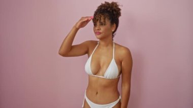 Bikinili genç bir kadın mutlu bir şekilde ayakta dikiliyor ve uzaklara bakıyor, izole edilmiş pembe arka planı güvenli bir şekilde araştırıyor.