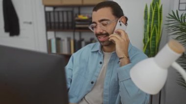 Ofis ortamında telefonla konuşan, gözlüklü ve mavi gömlekli İspanyol genç adam modern bir iş yerinde arka planda raflar ve bitkiler olan bir masada çalışıyor.