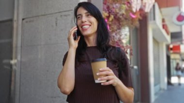 Güneşli bir günde telefonda konuşurken gülümseyen ve elinde kahve tutan bir kadın.