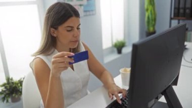 Parlak bir ofisteki bilgisayarı kullanarak elinde kredi kartı tutan kadın ciddi bir ifade sergiliyor ve muhtemelen finansal bir işlem üzerinde çalışıyor.