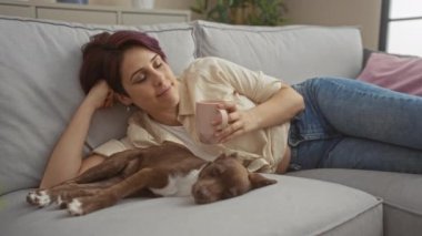 Oturma odasında genç, güzel bir kadın elinde kahve fincanı tutarken uykulu köpeğini nazikçe okşar, evde huzurlu bir neşe ve rahatlama anı yakalar..