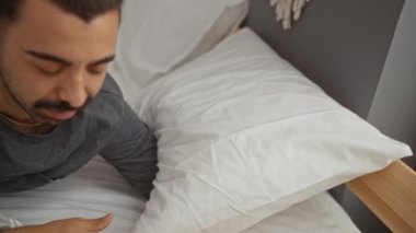 Gri gömlekli, bıyıklı, gözleri kapalı gülümseyen, rahat bir yatak odası ortamında beyaz bir yastık tutan, ev ortamında rahatlama ve rahatlama ima eden genç bir adam..