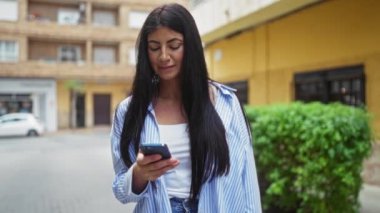 Esmer kadın akıllı telefonunu kullanarak bir şehir caddesinde duruyor, gülümsüyor ve dışarıda mutlu görünüyor..