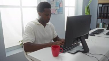 Modern ofis ortamında çalışan genç bir adam, masada kırmızı kahve fincanı ile bilgisayarda yazı yazan, profesyonel bir ortamda odaklanma ve başarı duygularını gösteren biri.