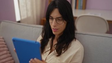 Oturma odasında elinde gözlüklerle tablet kullanan bir kadın rahat bir evde kitap okumaktan zevk alıyor..