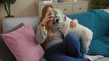 Kulaklık takan genç sarışın bir kadın oturma odasında köpeğiyle oturuyor, yastıklarla süslenmiş bir kanepede dinleniyor..