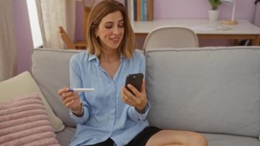 Pozitif hamilelik testi tutan kadın gülümsüyor ve oturma odasında akıllı telefona bakıyor.