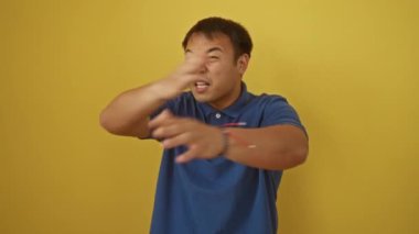 Kızgın genç Çinli adam ayakta, öfkeli ve hayal kırıklığına uğramış, izole edilmiş sarı arka planda öfke ve agresif bir ifadeyle bağırıyor.