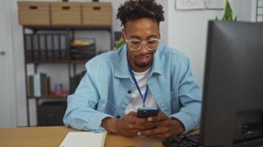 Ofisteki genç Afro-Amerikalı adam masasında ofis malzemeleri ve mobilyalarla çevrili bir akıllı telefon kullanarak gülümsüyor ve bunu kutluyor..