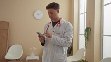 Genç İspanyol erkek doktor, bir klinik lobisinde, akıllı telefon kullanıyor, boynunda kırmızı steteskop olan beyaz bir ceket giyiyor, beyaz sandalyelerin ve saksı bitkilerinin yanında duruyor..