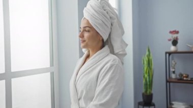 Kafasında bez bornoz ve havluyla spa 'da oturan İspanyol bir kadın, sakin bir iç mekan güzellik merkezinde pencereden dışarı bakıyor.