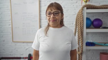 Beyaz tişörtlü, spor salonunda spor salonunda spor aletleri ve arka planda beyaz bir tahtayla kol kaslarını esneten kendinden emin, olgun bir İspanyol kadın..